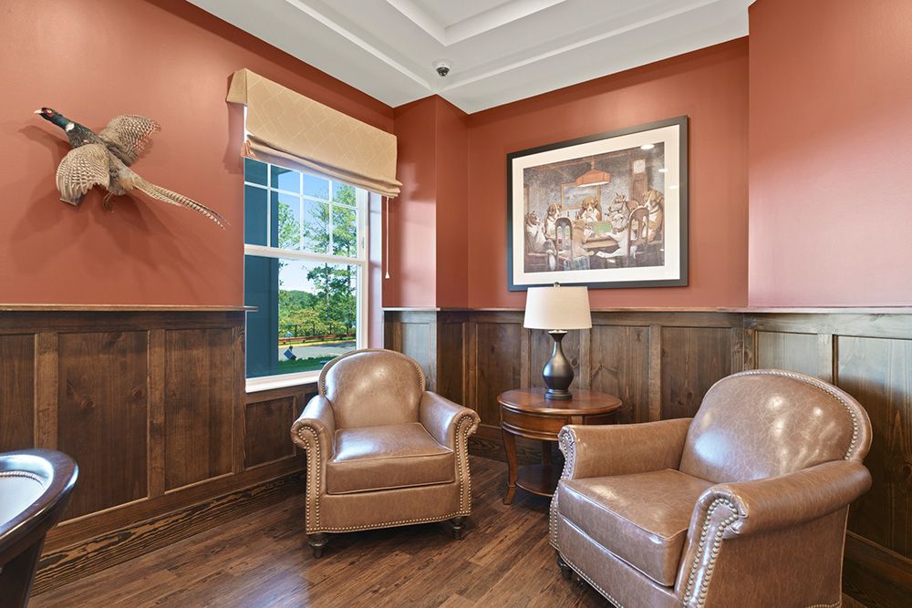 A cozy sitting area with leather armchairs and rich wood paneling, featuring a window, wall art, and a lamp on a side table.