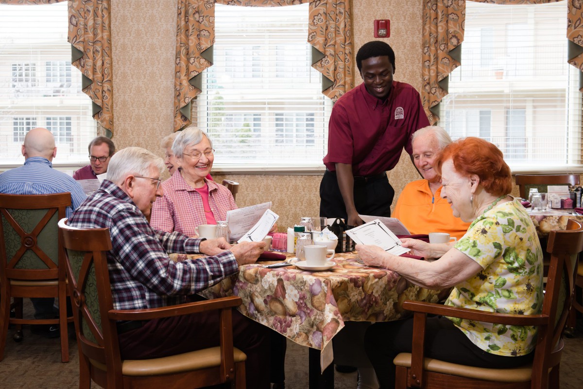 A group of senior citizens enjoying a meal together in a care home, assisted by a staff member.