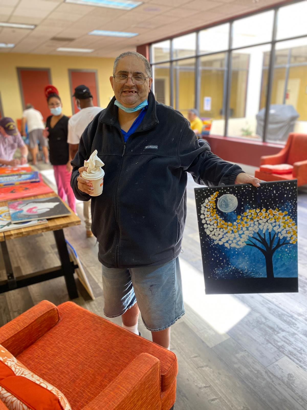A person proudly displays a painting they created while holding a cup inside an art studio.