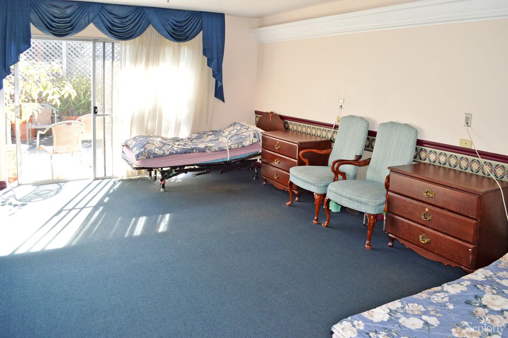 A spacious bedroom with sunlight streaming through the window, featuring a bed, multiple chairs, and elegant drapes.