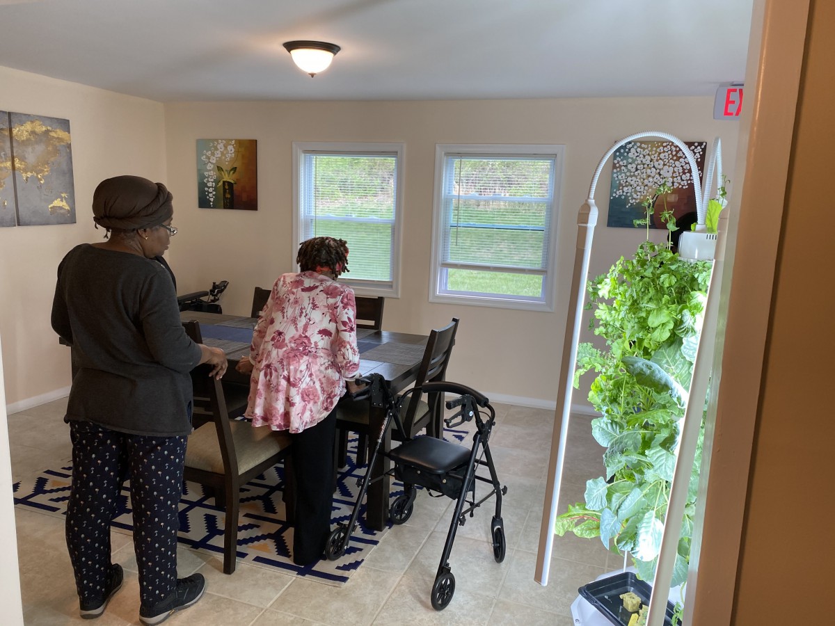 Two individuals standing in a dining area with a walker and indoor plants visible.