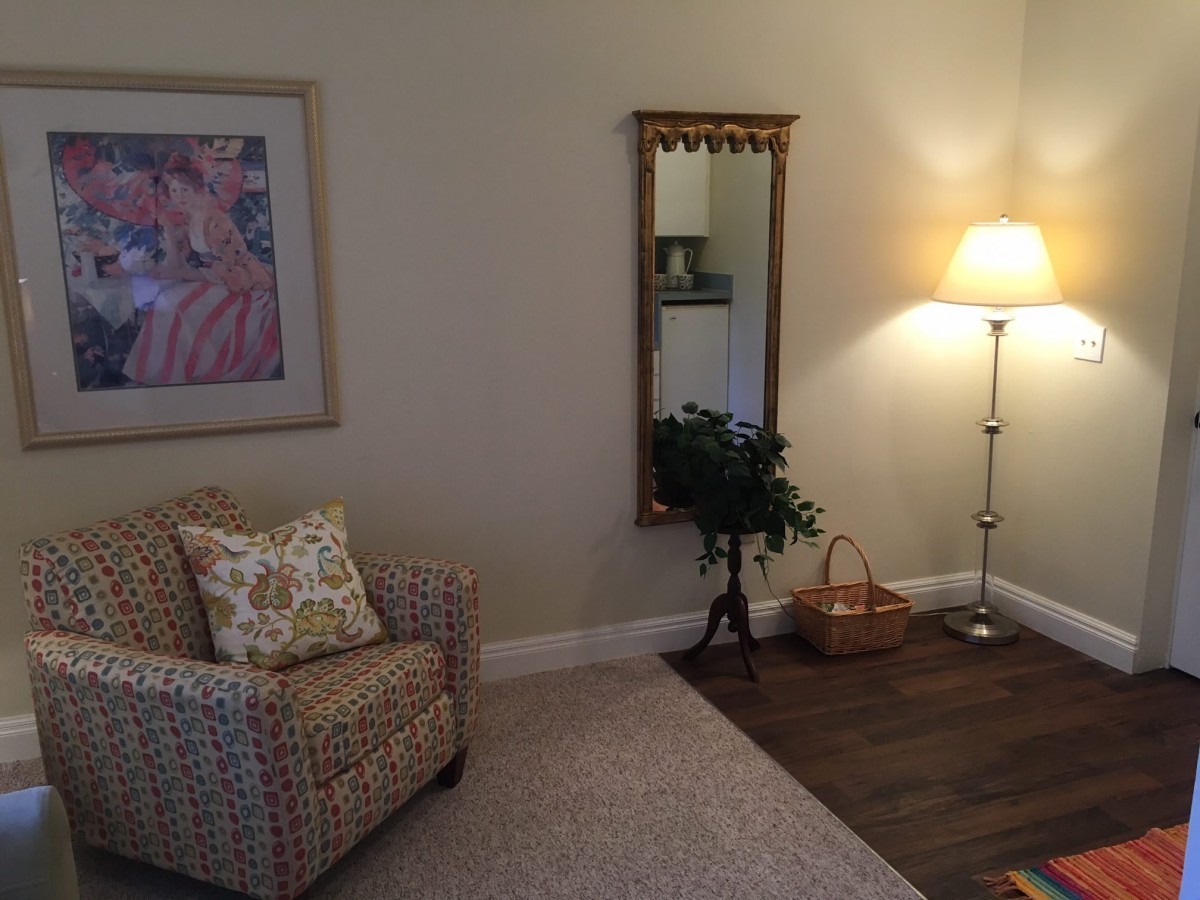 A cozy living room corner featuring a patterned chair, floor lamp, decorative mirror, artwork on the wall, and a small plant.