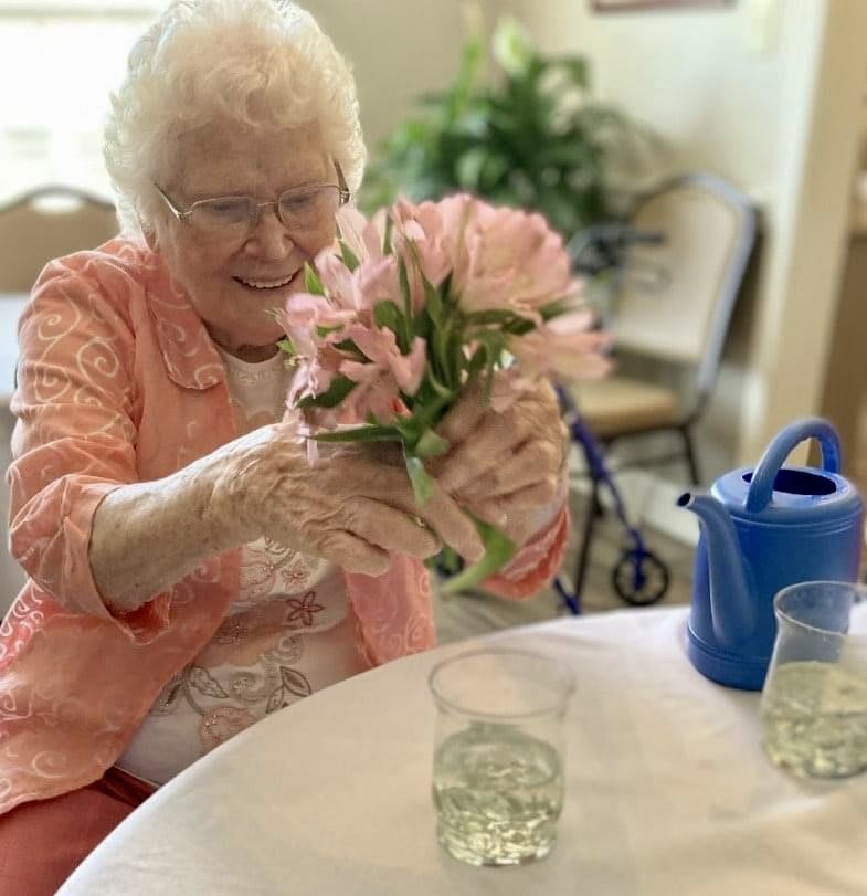 An elderly woman arranging flowers at a table with a watering can.