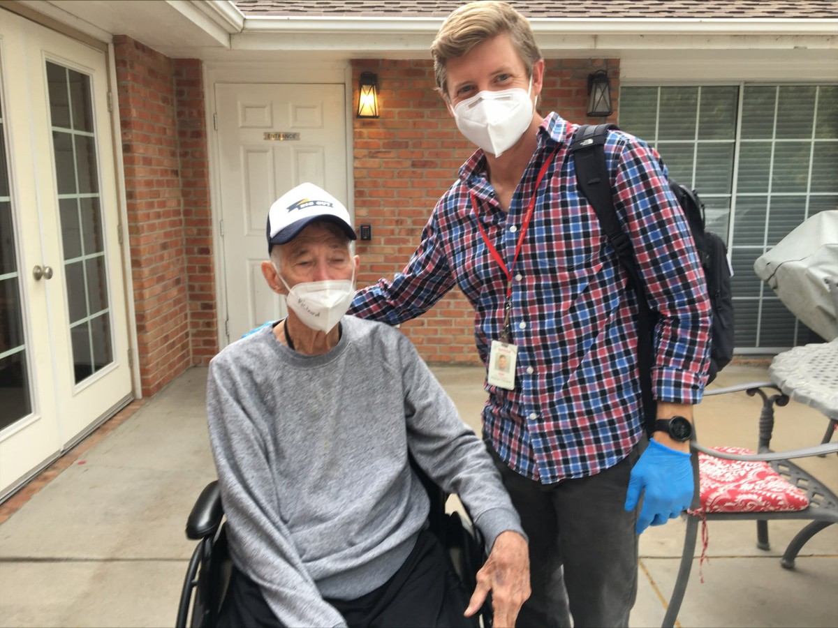 Two individuals wearing face masks are outside, one seated in a wheelchair and the other standing beside them.