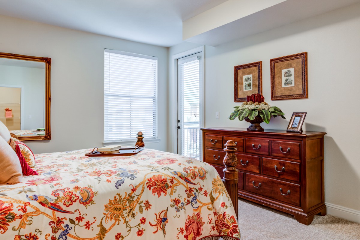 A cozy bedroom featuring floral bedding and a wooden dresser with decorative frames and a plant.