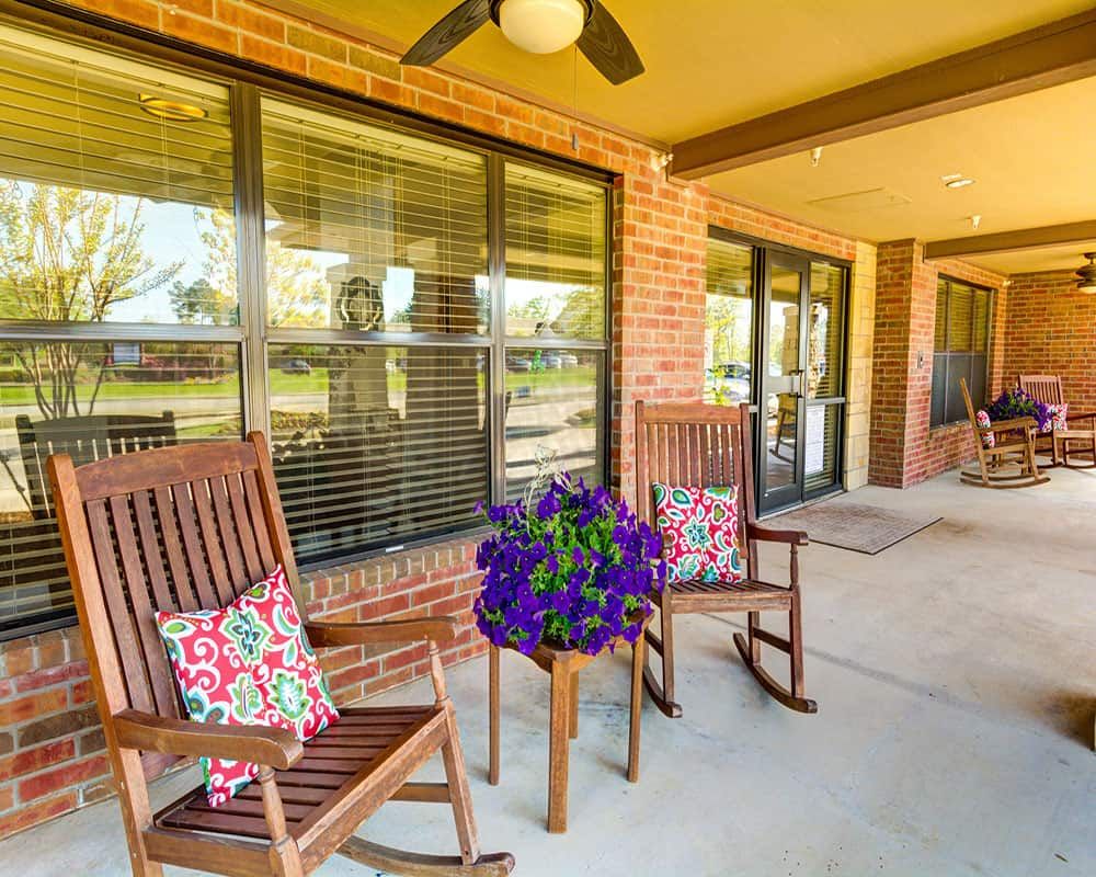 A cozy porch featuring two wooden rocking chairs with colorful cushions, a small table adorned with vibrant flowers, and large windows set into a brick wall.
