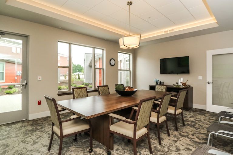 A modern conference room with a large table, several chairs, windows, and a wall-mounted television.