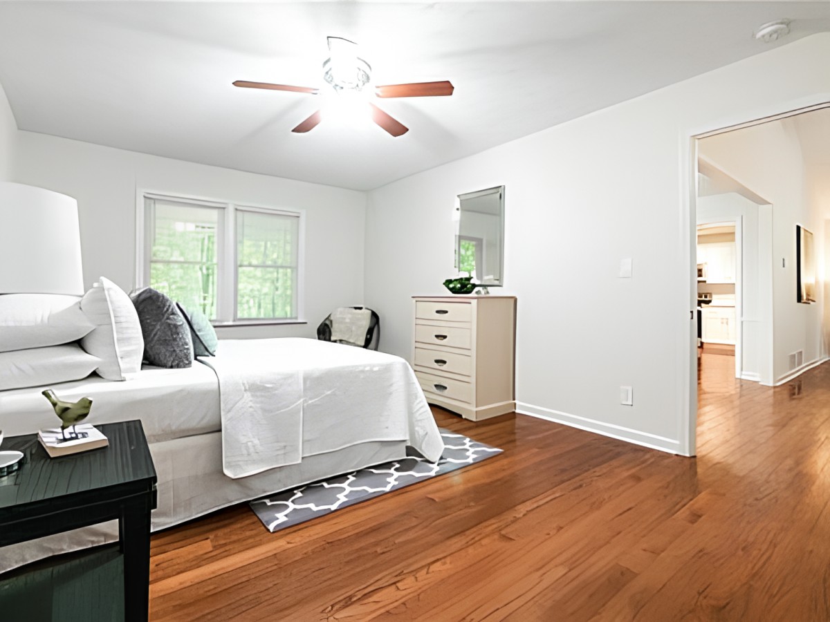 A bright and spacious bedroom with hardwood floors, featuring a neatly made bed, a dresser, and a ceiling fan.