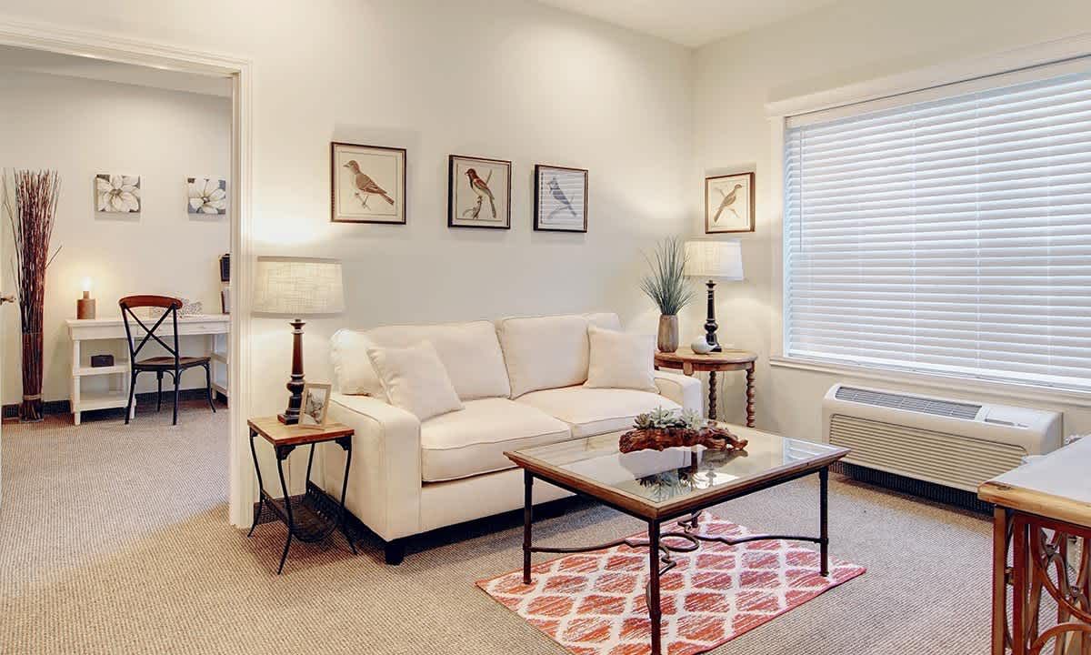 A cozy living room with a beige sofa, decorative pillows, and a wooden coffee table on a patterned rug. The walls are adorned with framed bird illustrations and two matching lamps provide warm lighting.