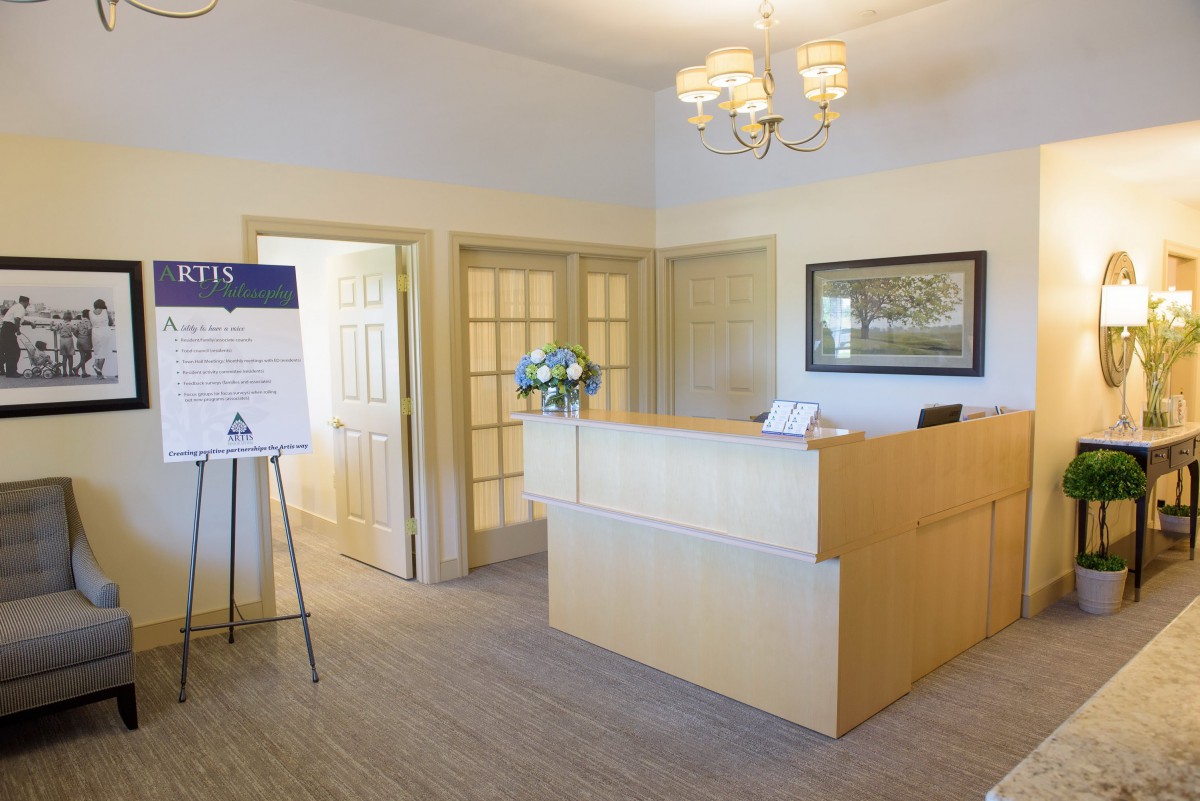 A bright office reception area with a desk, chandelier, and a signboard near the entrance.