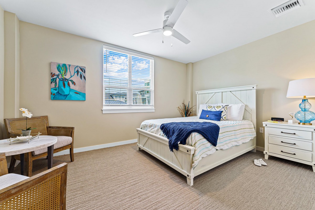 A cozy bedroom with a white bed, carpeted floor, and a ceiling fan under natural light from the window.