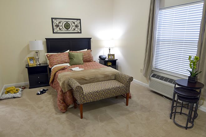 A cozy bedroom with a neatly made bed, decorative pillows, and a small bench at the foot. A lamp sits on the nightstand beside the bed.