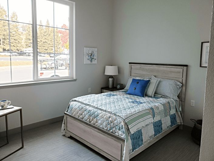 A cozy bedroom with a neatly made bed, featuring a patterned quilt and pillows, next to a window and a nightstand with a lamp.