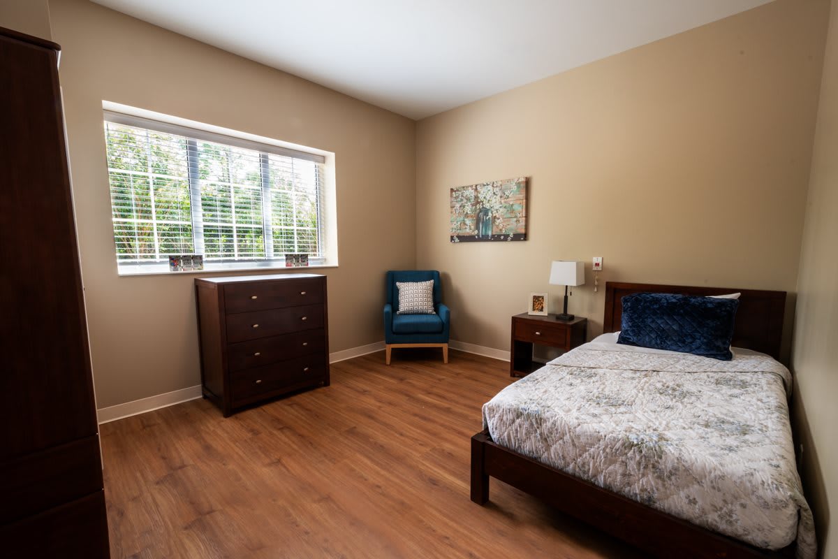 A cozy bedroom with wooden flooring, featuring a bed, a dresser, and a chair next to a window.