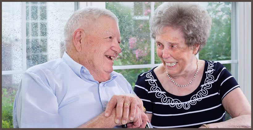 An elderly couple smiling and holding hands by a window.