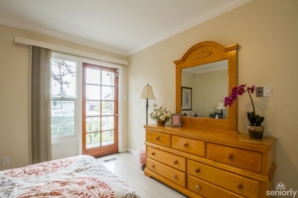 A cozy bedroom featuring a wooden dresser with a mirror, adorned with decorative flowers and illuminated by natural light from French doors.