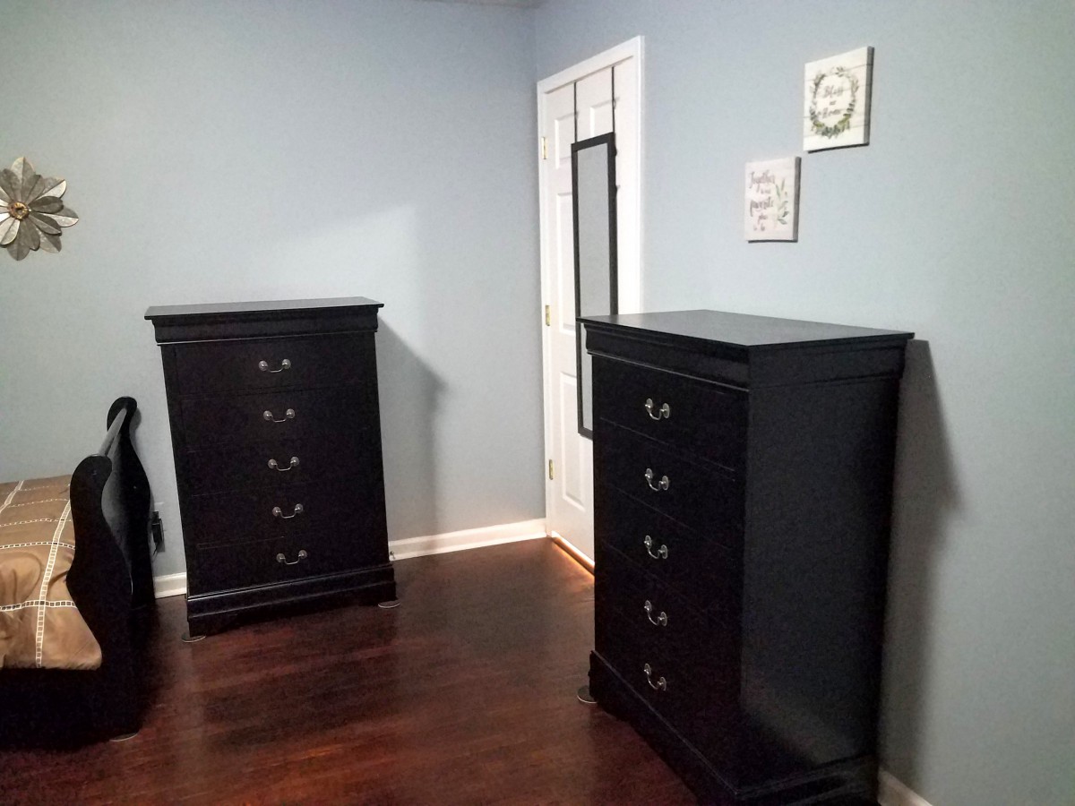 A neatly arranged bedroom with two black dressers, a mirror on the door, and wall art in a room with wooden flooring.