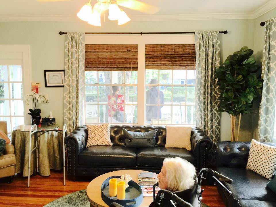 A cozy living room with a black leather sofa, large windows with curtains, and a ceiling fan above.