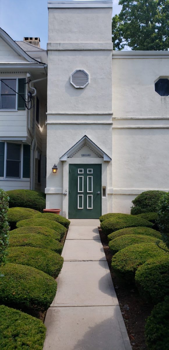 A building entrance with a green door and neatly trimmed shrubbery lining the pathway.