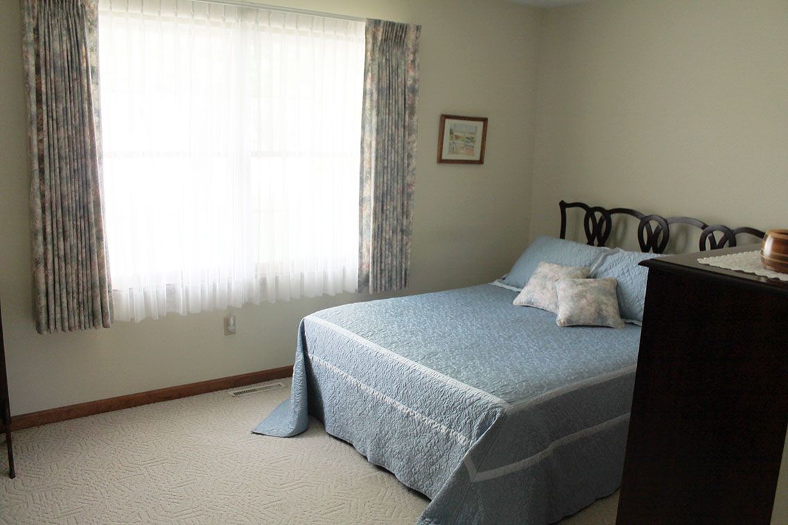 A simple bedroom with a blue bedspread and natural light coming through a window.