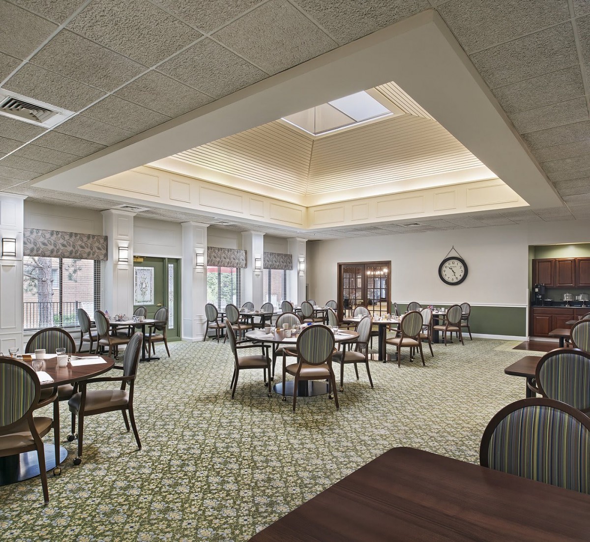 A spacious dining room with multiple tables and chairs, featuring carpeted flooring and large windows that let in natural light.