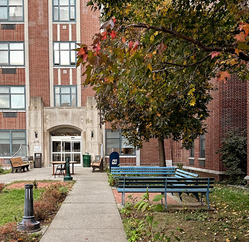The entrance of a brick building surrounded by trees and benches, featuring a well-maintained pathway.