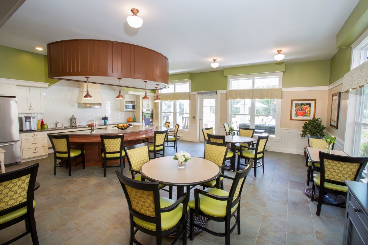 A spacious and well-lit dining area with round tables and cushioned chairs near an open kitchen.