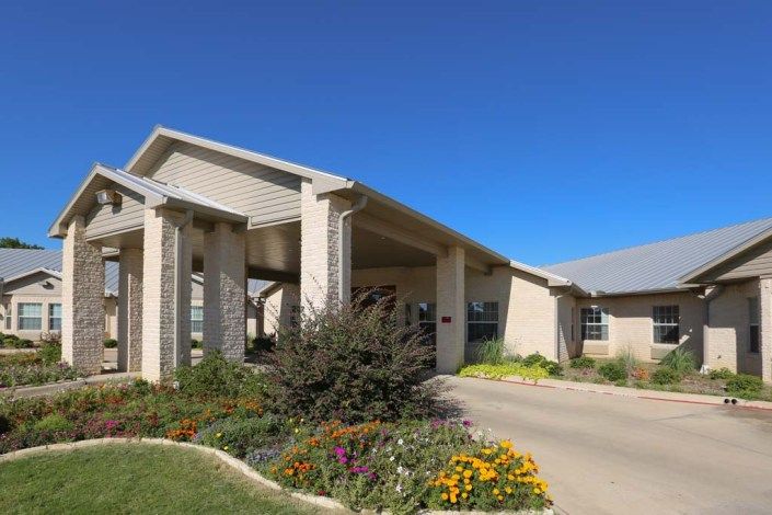 A modern building with a covered entrance, surrounded by landscaped gardens and a driveway.