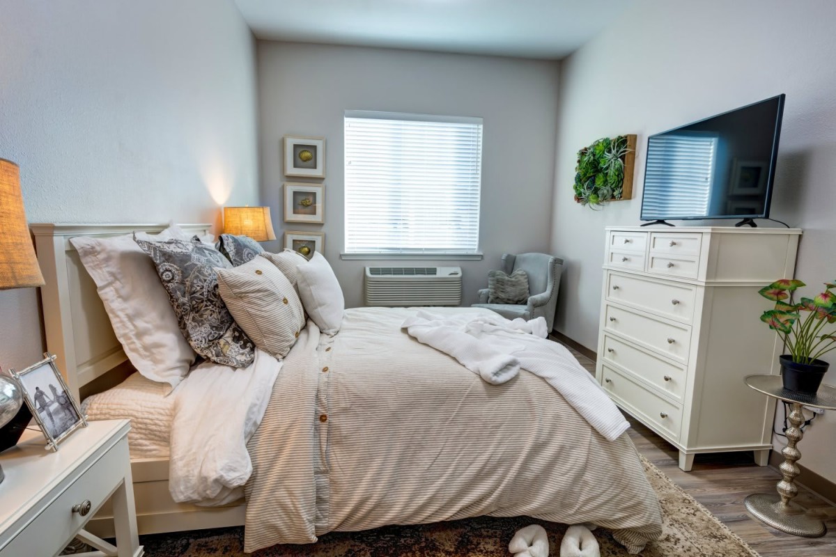 A cozy bedroom with a neatly made bed adorned with pillows, a bedside lamp, and natural light streaming in through the window.