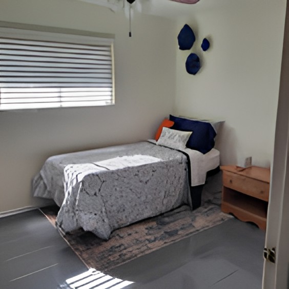 A cozy minimalist bedroom with a single bed, decorative pillows, and a nightstand under window blinds.