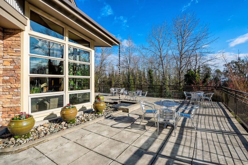 A modern patio with glass windows and outdoor seating, surrounded by trees on a sunny day.