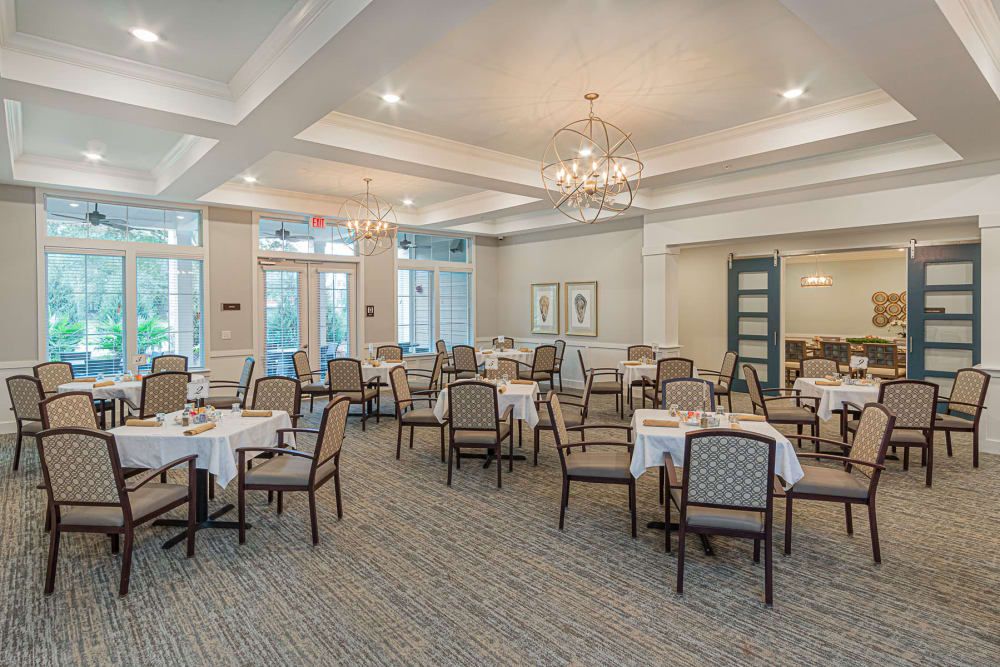 A spacious and elegantly decorated dining room with multiple tables and chairs, featuring a chandelier and large windows.