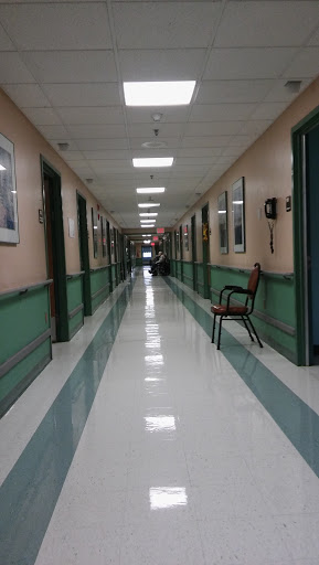 A quiet corridor in a hospital, featuring green and beige walls with photographs and a solitary chair.