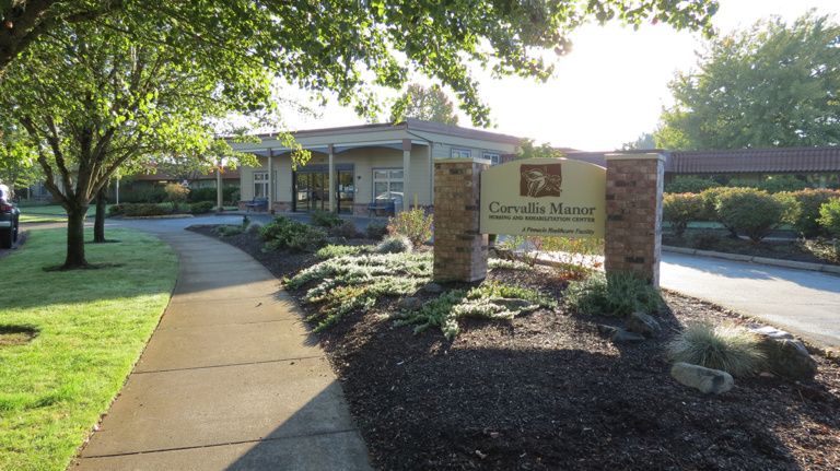 A view of a building surrounded by landscaping with a prominent sign and sidewalk under sunny conditions.