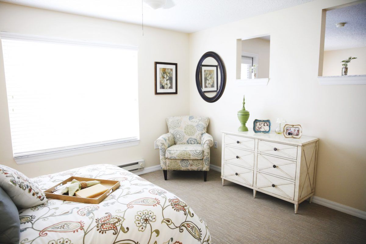 A bright and cozy bedroom with neutral tones, featuring an armchair, a dresser, and decorative artwork.