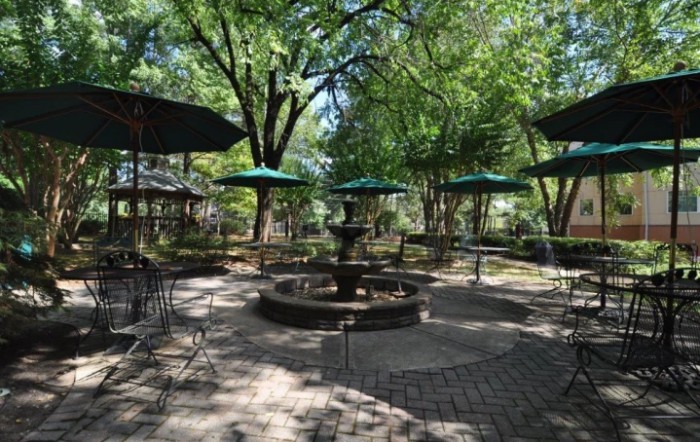 A tranquil outdoor patio with a central fountain, surrounded by tables and chairs under green umbrellas and shaded by tall trees.