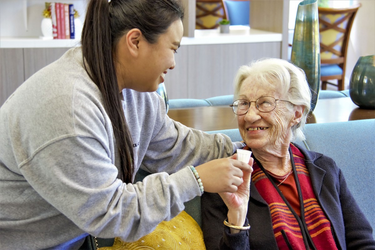 A caregiver assists an elderly woman, both smiling warmly.