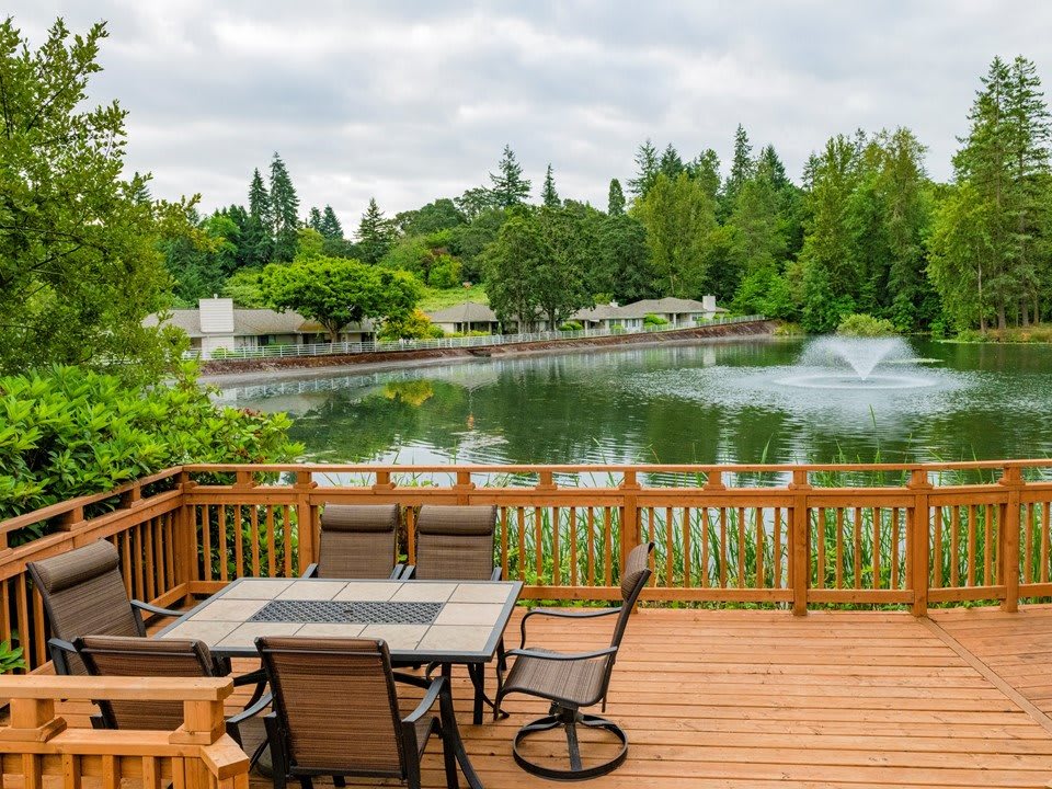 A wooden deck overlooks a serene lake with a fountain, surrounded by lush greenery.