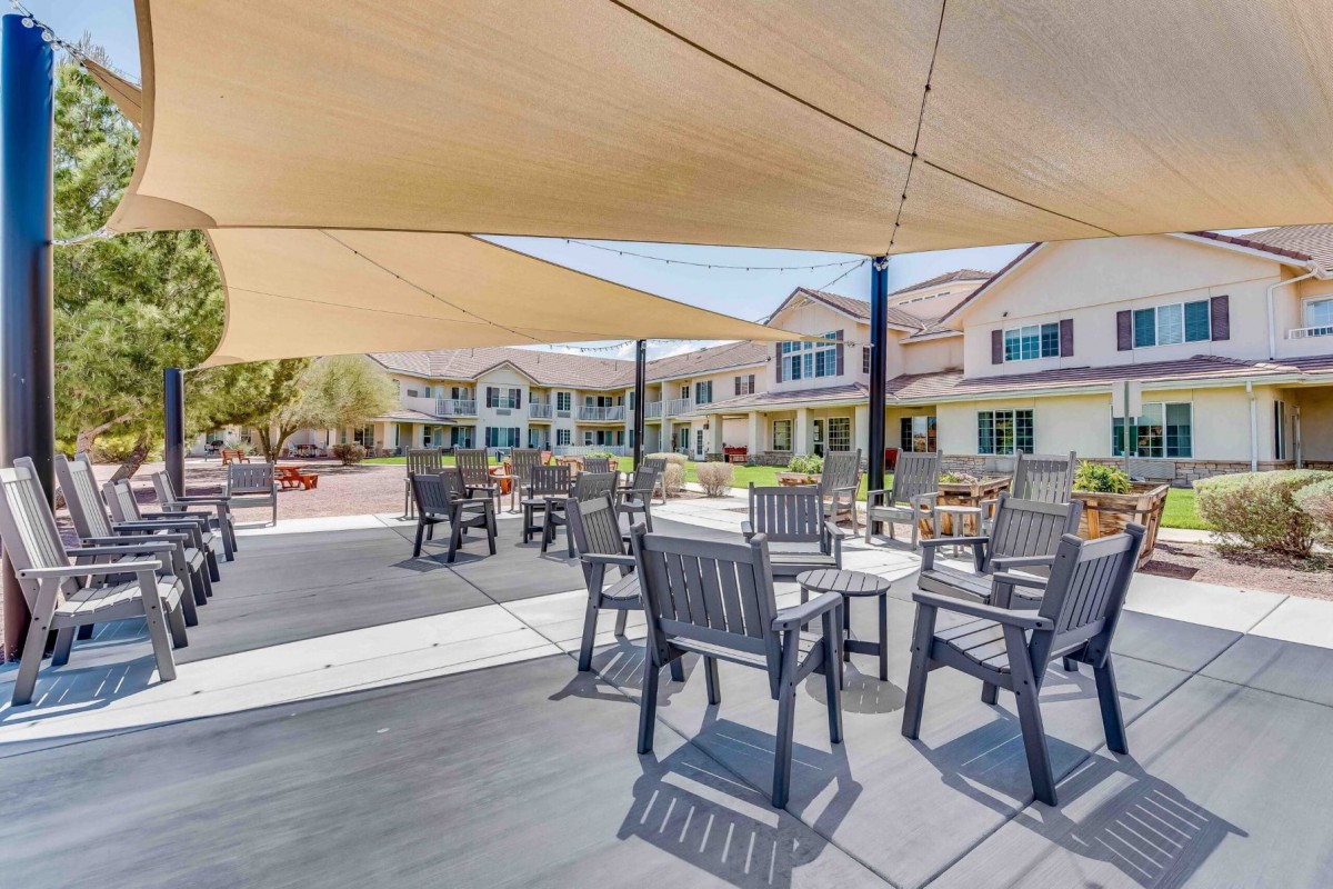 A shaded outdoor patio area with tables and chairs, adjacent to a large building on a sunny day.