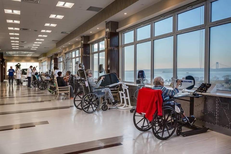 Elderly residents in a nursing home sitting by large windows enjoying the sunlight.