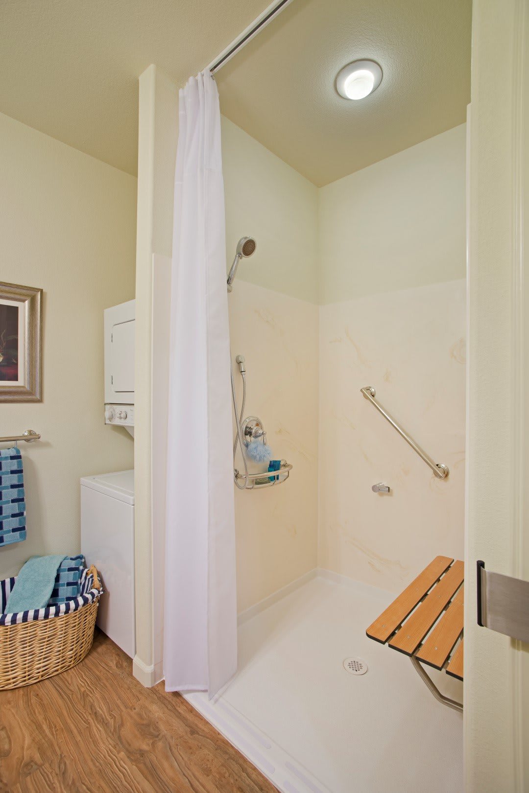 An accessible bathroom featuring a shower with grab bars, a curtain, and a bamboo bench, accompanied by neatly arranged towels and a laundry basket.