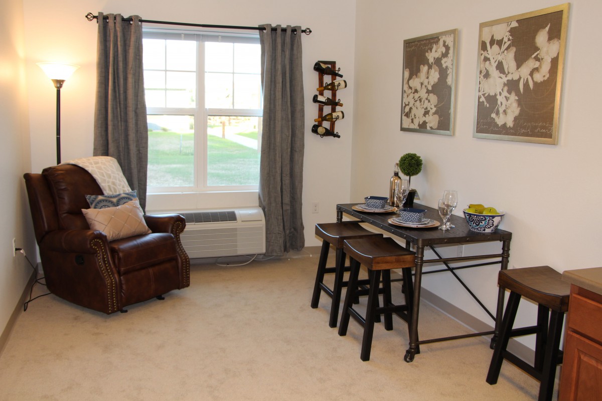 A cozy living and dining area with a large window, featuring comfortable seating, decorative artwork, and a neatly set table.