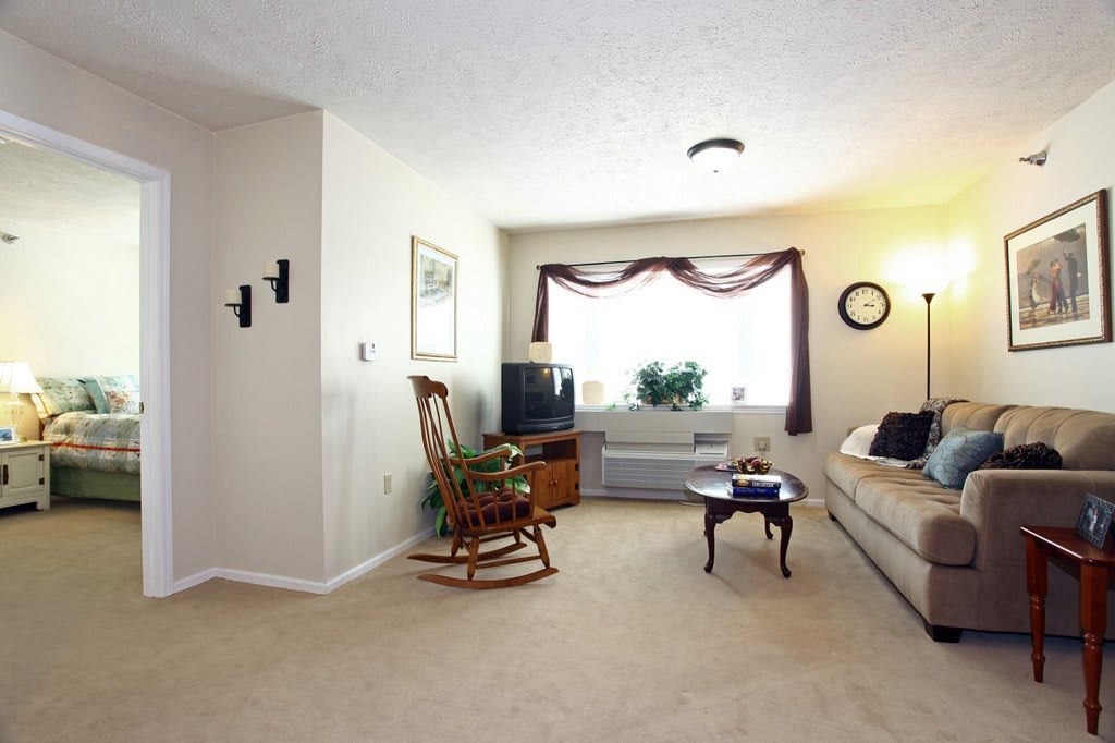 A cozy living room with a sofa, rocking chair, and coffee table facing a television under a bright window.