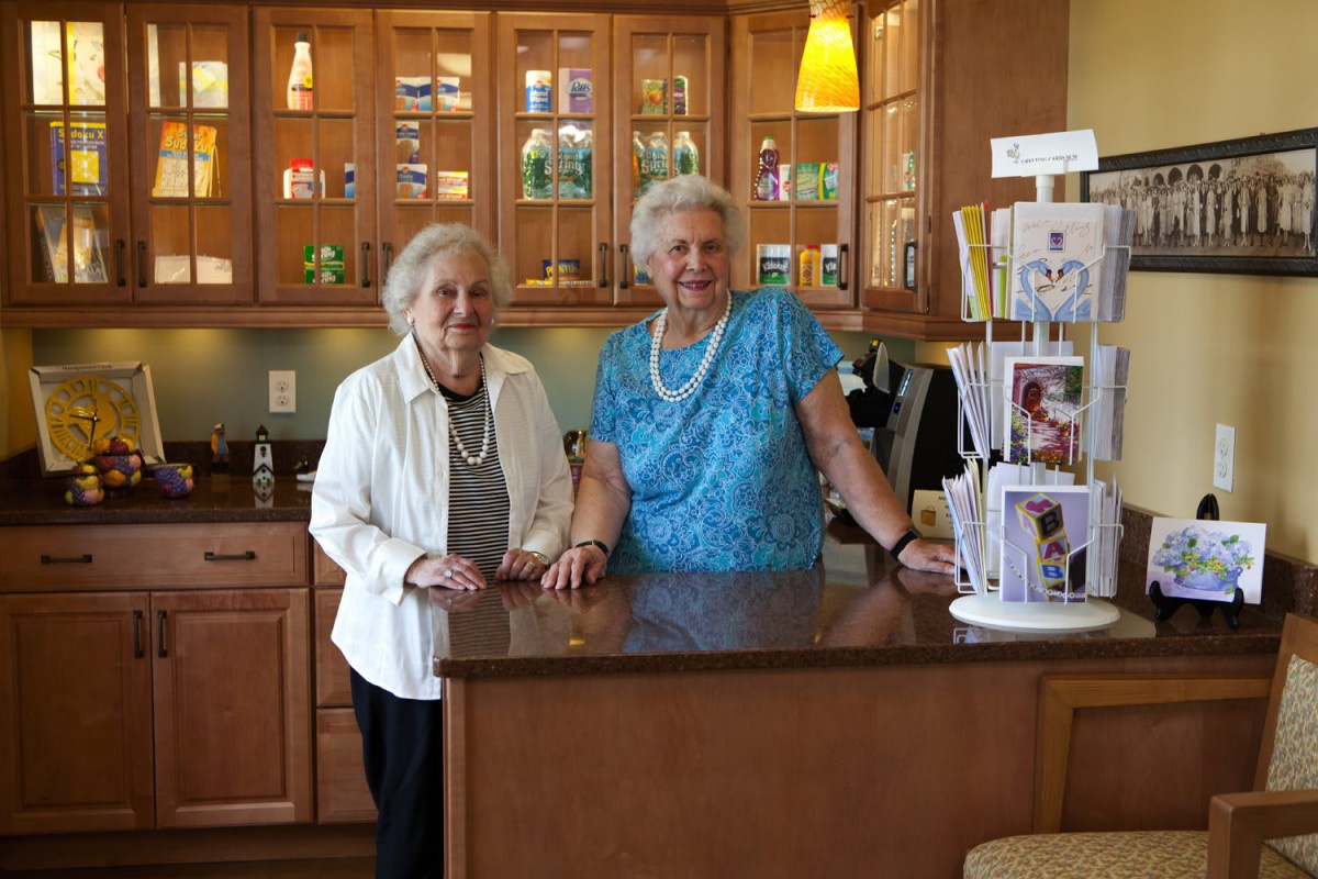 Two elderly women standing in a kitchen next to a display of greeting cards, smiling at the camera.