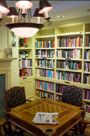 A cozy library room with a large bookshelf, a table with chairs, and a fireplace.