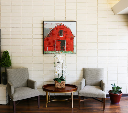 A cozy seating area featuring a red barn painting, two comfortable chairs, and a round table adorned with greenery.