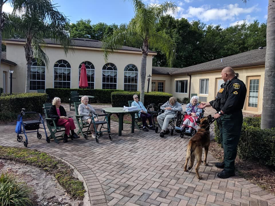 A group of elderly residents enjoys a visit from a police officer and his dog on the patio of a nursing home.