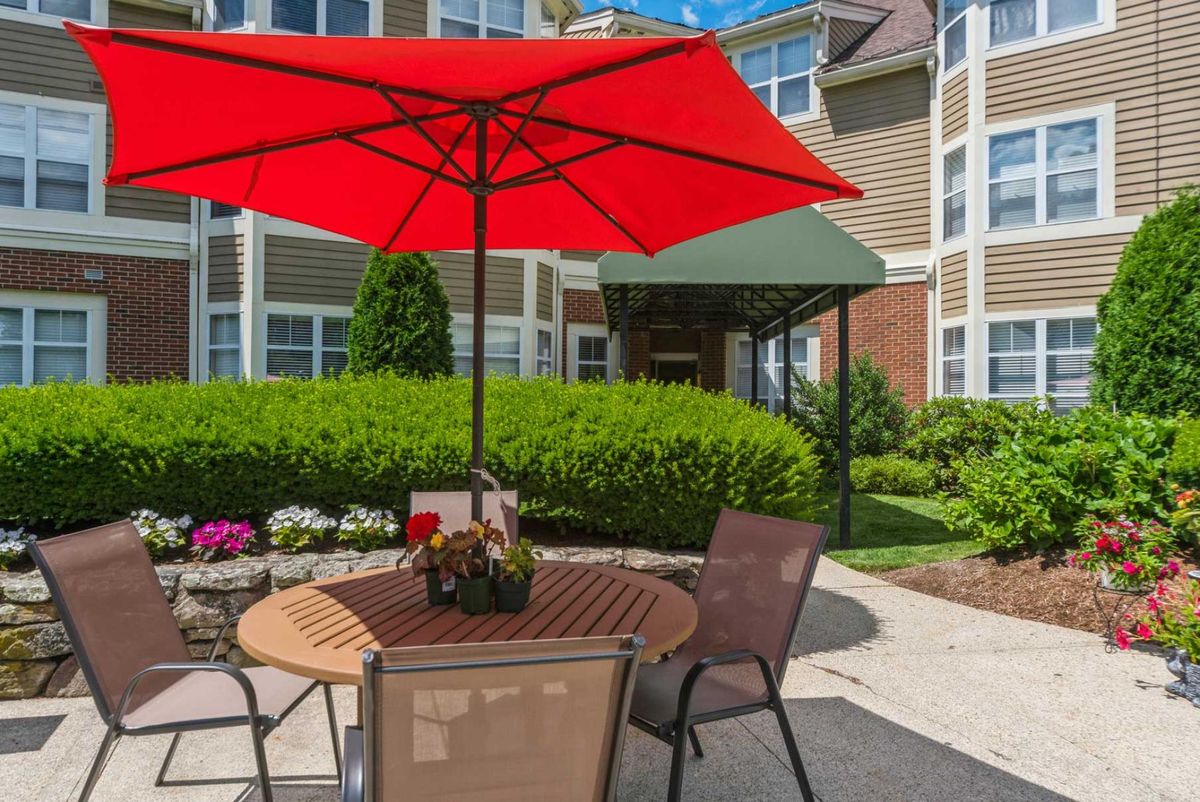 A cozy outdoor patio with a red umbrella, surrounded by lush greenery and flowers.