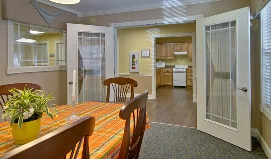 A cozy dining room with a table and chairs, leading into a kitchen through open French doors.