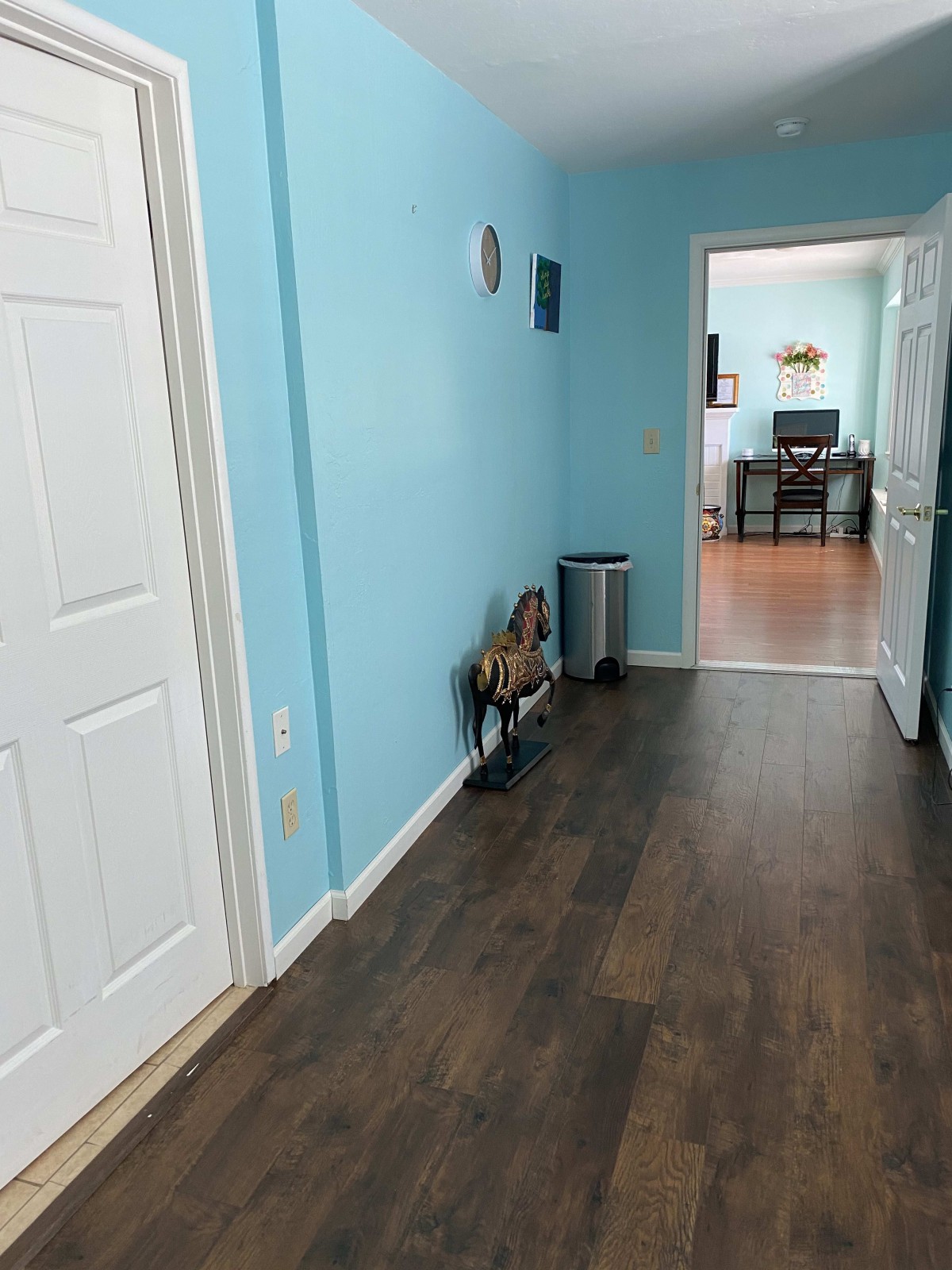 A hallway with blue walls and wood flooring, leading to another room with a desk.